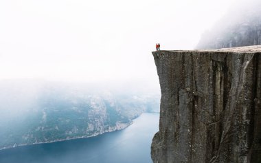 Preikestolen 'in kıyısında iki maceraperest yürüyüşçü duruyor. Norveç' te görkemli bir uçurum. Etrafı sisle çevrili, aşağıdaki fiyortun ve engebeli arazinin dramatik panoramik manzarasından hoşlanırlar..
