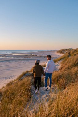 Bir çift gün batımında Danimarka 'da pitoresk bir plajın kumlu sahillerinde el ele yürür. Altın gökyüzü sakin suları yansıtıyor, sakin bir atmosfer yaratıyor romantik anlar için mükemmel..