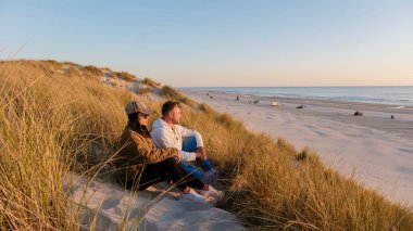 Danimarka 'da kumlu bir kumulda güneşin ufukta batışının tadını çıkaran bir çift. Yumuşak dalgalar kıyıya vurur, yansıma için mükemmel barışçıl bir atmosfer yaratır..