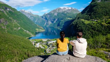 Çift Norveç 'te Geirangerfjord' a bakan bir kayanın üzerinde oturuyor. Etrafı muhteşem fiyort manzaraları ve yüksek dağlarla çevrili. Açık mavi gökyüzü ve yemyeşil, nefes kesici bir manzara yaratır..