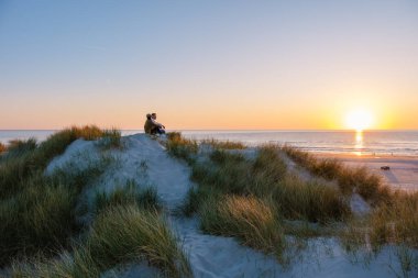 Bir çift uzun otlarla çevrili kumlu bir kum tepesinde oturur ve ufukta güneşin batışını izlerler. Vejers Strand Sahili Danimarka