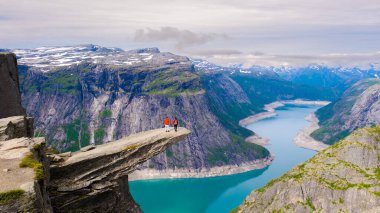 Norveç, Trolltunga 'da iki yürüyüşçü aşağıdaki turkuaz fiyortun nefes kesici manzarasını seyrederek dramatik bir uçurumun kenarında duruyor. Görkemli dağlar bulutlu bir gökyüzünün altındaki büyüleyici manzarayı sarar..