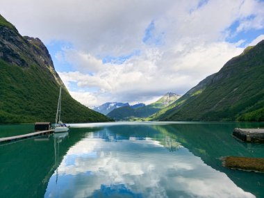 Norveç fiyordunun sakin suları sakin körfezi çevreleyen dramatik zirveleri ve canlı yeşilliği yansıtır. Bir yelkenli demir atmış, bu manzarada maceraya davetiye çıkarıyor..