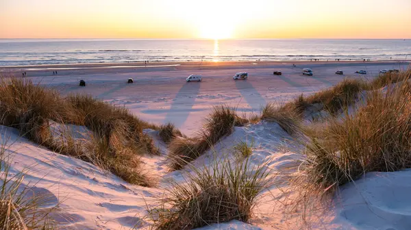 Altın gün batımı, Danimarka 'nın Vejers Strand sahilindeki kumlu bir plajda ılık bir parıltı yayar. Ziyaretçiler, park halindeki araçların kıyı şeridini noktaladığı akşamın keyfini çıkarıyorlar. 