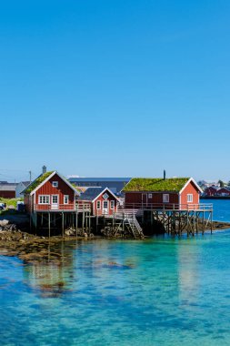 Parlak kırmızı evler, Lofoten Norveç 'in sakin turkuaz sularıyla çevrili cambaz ayaklıkları üzerinde duruyor. Açık gökyüzü ve sakin deniz, Norveç 'in doğal güzelliklerini gözler önüne sermek için mükemmel bir manzara yaratır..