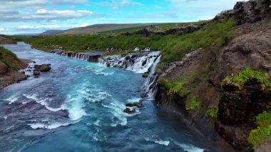 İzlanda Barnafoss ve Hraunfossar 'ı keşfedin, engebeli arazide çağlayan güzel şelaleler, yemyeşil ve açık mavi gökyüzü ile çevrili..