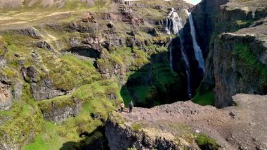 İki yürüyüşçü, İzlanda 'daki yeşil kayalıklardan aşağı çağlayan etkileyici bir şelaleye bakan kayalık bir uçurumda duruyor. Bu uzak yerin görkemli doğal güzelliği, Glymur şelale kanyonu İzlanda