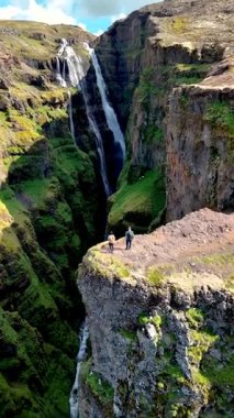 Göz kamaştırıcı bir şelale, İzlanda 'nın yemyeşil manzaralarıyla çevrili sarp kayalıklardan aşağı akıyor. Ziyaretçiler, İzlanda 'daki doğal güzellik, Glymur şelalesi kanyonu tarafından korkuyla kanyonların kenarında dururlar.