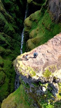 Maceracılar, canlı yeşil yosunlarla çevrili dar bir kanyona bakan nefes kesici bir uçurumun kenarında dururlar. Güneş ışığı İzlanda 'daki dramatik araziyi aydınlatır, Glymur şelale kanyonu.