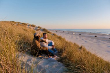 İki arkadaş, uzun çimlerle çevrili kumlu bir kumulda, Vejers Strand sahilindeki sakin plajda gün batımını seyrederken rahatlar..