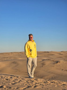 Bir gezgin, geniş kumlu tepelerin ortasında parlak sarı bir kapüşonlu giysiyle Danimarka 'nın çarpıcı manzarasının tadını çıkarıyor. Açık mavi gökyüzü, sahnenin sakin ve maceraperest atmosferini arttırır..