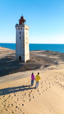 İki kişi kumlu bir kıyı şeridi boyunca el ele yürürken, tarihi bir deniz fenerine hayran kalarak sıcak güneşin tadını çıkarıyorlar. Sakin okyanus dalgaları bu huzurlu Danimarka manzarasını tamamlıyor. Rubjerg Knude