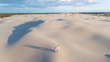 Danimarka 'nın geniş, dalgalı beyaz kum tepelerinde el ele dolaşan iki birey. Günbatımının sıcak altın ışığı uzun gölgeler bırakır Rabjerg Mile Dunes, Danimarka.