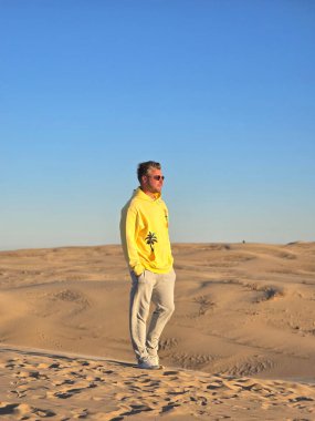 Genç bir adam, Danimarka 'daki altın kum tepelerinde emin adımlarla güneşin altında güneşleniyor. Açık mavi gökyüzü, sakin atmosferi arttırır. Açık hava maceraları için mükemmeldir..