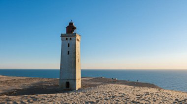 Rubjerg Knude deniz feneri kuleleri kumlu kumulların üzerinde, güneş batarken Kuzey Denizi manzaralı. Ziyaretçiler, Danimarka kıyı şeridinin dingin güzelliğinin keyfini çıkararak sakin bir atmosfer yaratıyorlar..