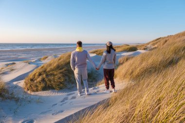 Danimarka kıyısındaki altın kum tepelerinde el ele gezen bir çift. Uzun otlar ve nefes kesici okyanus manzaralarıyla çevrili. Blokhus Strand, Danimarka