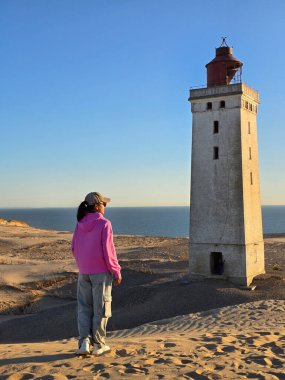 Tarihi Rubjerg Knude deniz fenerine bakan bir ziyaretçi, çarpıcı bir sahilin arka planına bakıyor. Altın saat güneşi, keşif ve fotoğrafçılık için mükemmel sakin bir atmosfer yaratır..
