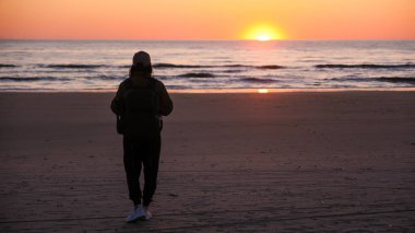 Danimarka 'da güneş ufukta batarken kumlu sahil boyunca yürüyen yalnız bir figür, yumuşak dalgalar ve gökyüzünü kucaklayan sıcak renklerle sakin bir atmosfer yaratıyor..