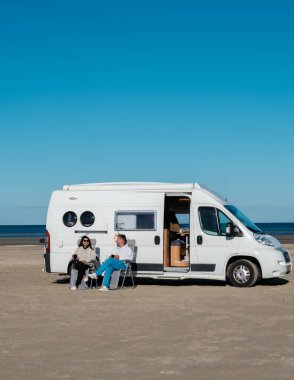 Çiftler Lakolk Strand, Romo Danimarka 'da kumlu bir plajda dinleniyorlar ve sakin okyanus manzarasının tadını çıkarıyorlar. Konforlu karavanları yakınlarda duruyor, doğa yolculuklarının ve açık hava maceralarının ruhunu somutlaştırıyor..