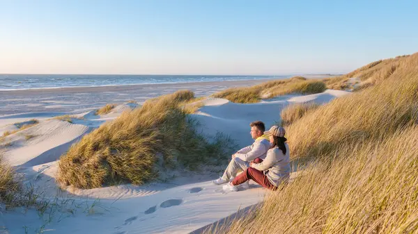 Güneş, Blokhus Strand, Danimarka 'daki sakin plajın üzerinde batarken, bir çift, yemyeşil çimlerle çevrili sıcak kum tepelerinde oturup, huzurlu anın tadını çıkarıyorlar..