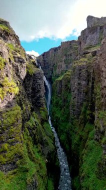 Parlak bir şelale, İzlanda 'daki Glymur şelalesi kanyonunda yemyeşil yeşilliklerle çevrili engebeli uçurumlardan aşağı yuvarlanır. Parlak gökyüzü bu nefes kesici doğa harikasına dramatik bir dokunuş ekler..