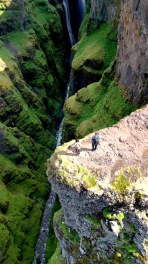 Hava manzarası, İzlanda 'daki görkemli bir kanyonun kenarında duran maceraperestlerin canlı yeşil uçurumlar ve aşağıda kıvrımlı bir nehir olan İzlanda' daki Glymur şelalesi kanyonunu göstermektedir.