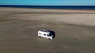 Romo Danimarka Lakolk plajının huzurlu manzarasını deneyin. Bir karavan yumuşak kumlu sahilde ilerliyor. Etrafı geniş bir kıyı şeridi ve açık mavi gökyüzüyle çevrili..
