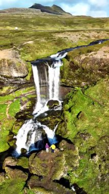 Sersemletici bir şelale İzlanda 'da yosun kaplı kayalara dökülüyor ve iki figür yemyeşil çevreyi keşfediyor..