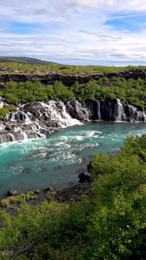 İzlanda 'da eşsiz bir şelale sistemi olan Hraunfossar' ın nefes kesici güzelliğini keşfedin. Kristal berrak sular, parlak mavi bir gökyüzünün altındaki canlı yeşil manzarayla çevrili volkanik kayaların üzerinde çağlar..