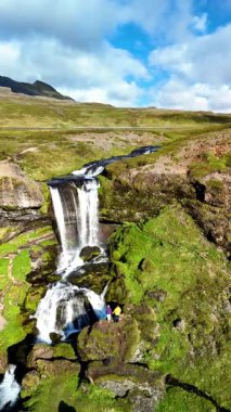 Ziyaretçiler, yosun kaplı kayalardan zarif bir şekilde akan Selvallafoss şelalesinin yanında huşu içinde duruyorlar. İzlanda 'nın nefes kesici manzarası İzlanda' da canlı yeşillik ve dramatik gökyüzünü sergiliyor