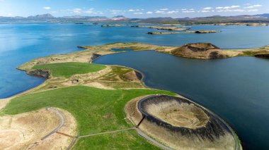 İzlanda 'daki Myvatn kraterlerinin nefes kesen güzelliğini deneyimlemek, berrak mavi gökyüzünün altında dingin göl yansımaları arasında eşsiz volkanik jeoloji sergiliyor. Doğa severler ve fotoğrafçılar seviniyor..