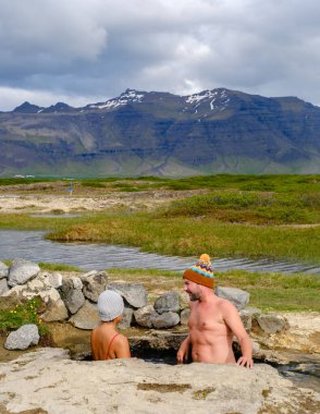 Two people enjoy a rejuvenating soak in the natural Landbrotalaug Hot Spring, set against the stunning backdrop of Icelands mountains and lush greenery.