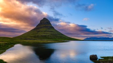 Kirkjufell Dağı rengarenk bir gökyüzüne çarpıcı bir şekilde yükselir. Yansıması aşağıdaki sakin sularda parıldar. Bu ikonik simge, İzlanda 'nın doğal güzelliklerini alacakaranlıkta yakalar..