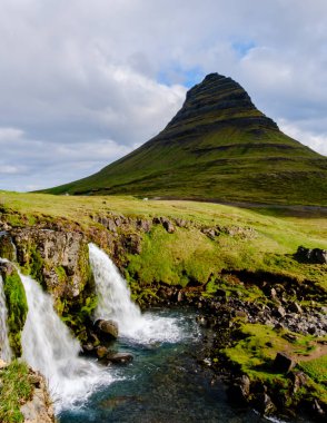 Nefes kesici Kirkjufell, yemyeşil yeşilliklerle birlikte dramatik bir şekilde yükseliyor. Yakınlarda, büyüleyici şelaleler huzurlu bir nehre dökülüp İzlanda 'nın güzelliklerini yansıtan pitoresk bir manzara yaratıyor..