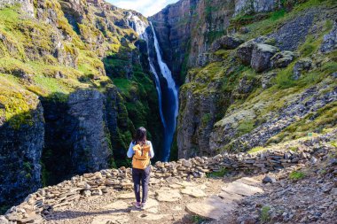Witness the majestic Glymur Waterfall cascading down cliffs in Icelands stunning landscapes. A hiker stands in awe, taking in the natural beauty that surrounds this unique location in the wilderness.