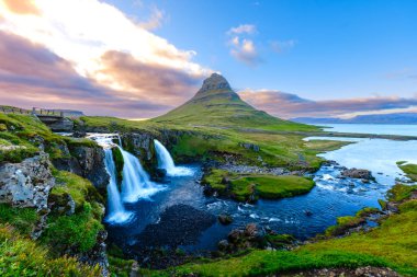 Kirkjufell dağ kuleleri, çağlayan şelaleler aşağıdaki sakin sulara hücum ederken yemyeşil tepelerin üzerindedir. Parlayan gökyüzü güneşin renkleriyle parıldıyor, büyüleyici bir atmosfer yaratıyor..