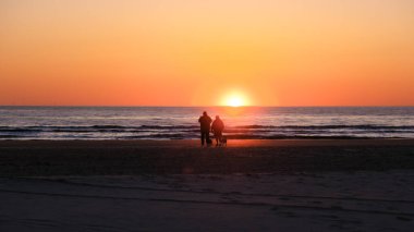 Danimarka 'nın Skagen kentindeki kumlu plajda, nefes kesici bir gün batımında köpeklerinin yanında yürüyen iki kişinin siluetleri gibi sakin bir an yaşanıyor. Gökyüzü sıcak turuncu ve pembe renklerle boyanmıştır..