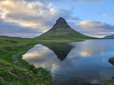 Kirkjufell dağı, aşağıdaki sakin sulara güzel bir şekilde yansıyan dramatik bulutlar zeminine karşı dimdik ayakta duruyor. Bu büyüleyici manzara İzlanda 'nın doğal güzelliğini yansıtıyor..