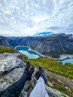 Two people enjoy a moment of tranquility while sitting on a rocky outcrop, overlooking a stunning fjord in Norway. The vast sky and lush mountains enhance the serene atmosphere of Trolltunga, Norway