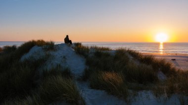 Huzurlu Danimarka plajının üzerinde altın güneş batıyor, kumlu sahile sıcak renkler döküyor ve huzurlu bir akşamın tadını çıkarıyor. Doğa ve rahatlama bu resim ortamında birleşiyor.