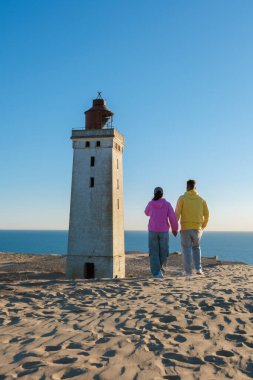 Bir çift, tarihi bir deniz feneri olan Rubjerg Knude 'un yanında el ele romantik bir gezinti yapar. Güneş ufukta batarken, kumlu arazi ve okyanus dalgaları üzerinde sıcak bir parıltı yayar..