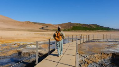A traveler explores the stunning Hverir geothermal area of Iceland, walking on a wooden path surrounded by colorful mineral-rich terrain and blue skies. Adventure awaits in this unique landscape.