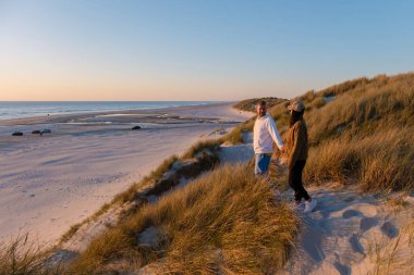 Gün batımında Danimarka 'nın sakin plaj manzarasının tadını çıkararak, el ele kumulların üzerinde birkaç gezinti. Güneş, dingin manzaraya ve okyanus dalgalarına sıcak altın bir ışık saçar..
