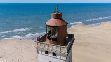 Ziyaretçiler, Kuzey Jutland Danimarka 'nın Rubjerg Knude sahillerine bakan tarihi deniz fenerini keşfediyorlar. Sakin okyanus berrak mavi gökyüzünün altında parıldıyor.