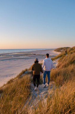 Gün batımında Danimarka 'nın Vejers Strand plajındaki sakin plajda el ele dolaşırlar. Yumuşak altın ışık sakin dalgaları yansıtır.
