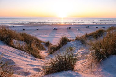 Büyüleyici bir günbatımı, Danimarka 'da kumlu bir plajın üzerinde canlı renkler bırakır. Kıyıda hafif dalgalar ve sahil şeridi boyunca park etmiş araçlar. Vejers Strand plajı.