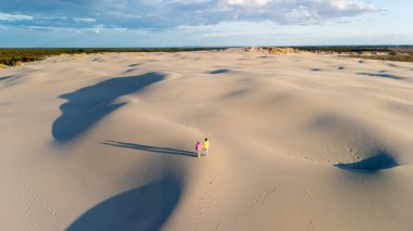 Danimarka 'nın büyüleyici manzarasında dolaşın. Yumuşak kum tepeciklerinin titrek bir gökyüzünün altında sonsuz bir şekilde uzandığı yerde. Kum tepeleri boyunca yürüyen iki figür, Rabjerg Mile Kumulları, Danimarka.