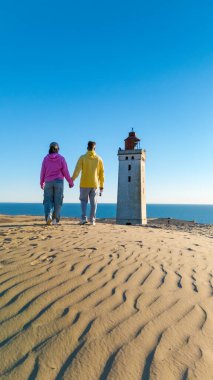 Arka planda tarihi bir deniz feneri olan kum tepeleri boyunca el ele tutuşan iki kişi. Açık mavi gökyüzü, elbiselerinin parlak rengini artırıyor, Rubjerg Knude deniz feneri Danimarka