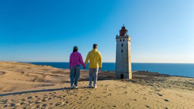 İki birey, denize bakan kumlu bir kumulda el ele dururlar. Arka planda açık mavi bir gökyüzünün altında tarihi bir deniz feneri vardır. Rubjerg Knude Deniz feneri Danimarka Jutland