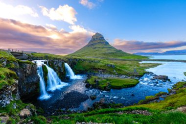 Kirkjufell dağ kuleleri İzlanda 'nın yemyeşil manzaraları üzerinde, yakındaki bir şelaleden çağlayarak akan su ile. Parlak renkler gün batımında gökyüzünü aydınlatır, dingin bir atmosfer yaratır..